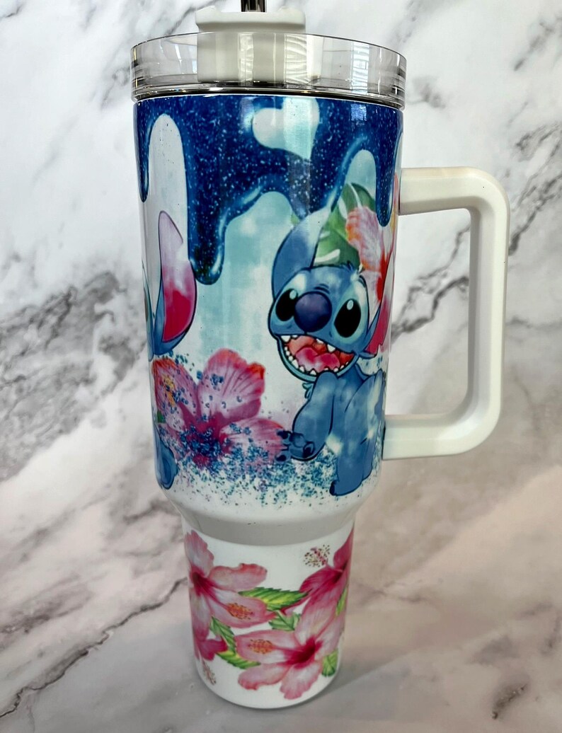 May include: White travel mug with a blue and white cartoon character design. The character is Stitch from Lilo & Stitch. The mug has a white handle and a clear lid. The design features pink flowers and blue glitter.
