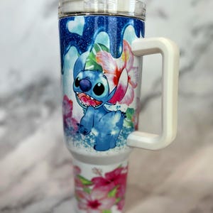 May include: A white stainless steel travel mug with a blue and white cartoon character design. The mug has a white handle and a clear lid. The design features a blue character with a pink flower and green leaves. The mug is on a white marble surface.