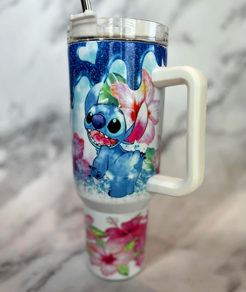 May include: A white stainless steel travel mug with a blue and white cartoon character design. The mug has a white handle and a clear lid. The design features a blue character with a pink flower and green leaves. The mug is on a white marble surface.