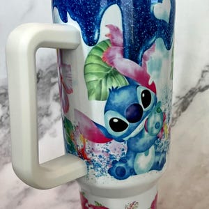 May include: A white stainless steel tumbler with a blue, pink, and green floral design featuring Stitch from Lilo & Stitch. The tumbler has a white handle and a clear lid with a straw.