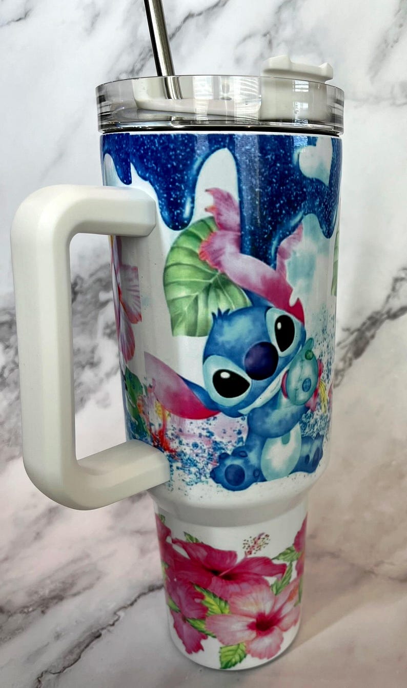 May include: A white stainless steel tumbler with a blue, pink, and green floral design featuring Stitch from Lilo & Stitch. The tumbler has a white handle and a clear lid with a straw.