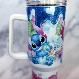 May include: A white stainless steel travel mug with a blue and pink floral design featuring Stitch from Lilo & Stitch. The mug has a white handle and a clear lid with a straw.