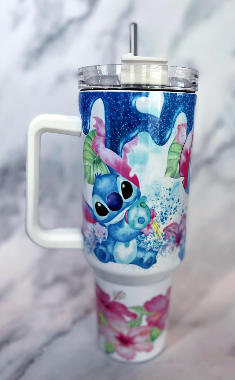 May include: A white stainless steel travel mug with a blue and pink floral design featuring Stitch from Lilo & Stitch. The mug has a white handle and a clear lid with a straw.