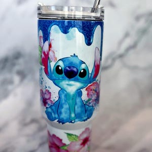May include: A white stainless steel tumbler with a blue and white watercolor design of Stitch from Lilo & Stitch. The tumbler has a clear lid and a stainless steel straw. The design also includes pink hibiscus flowers.