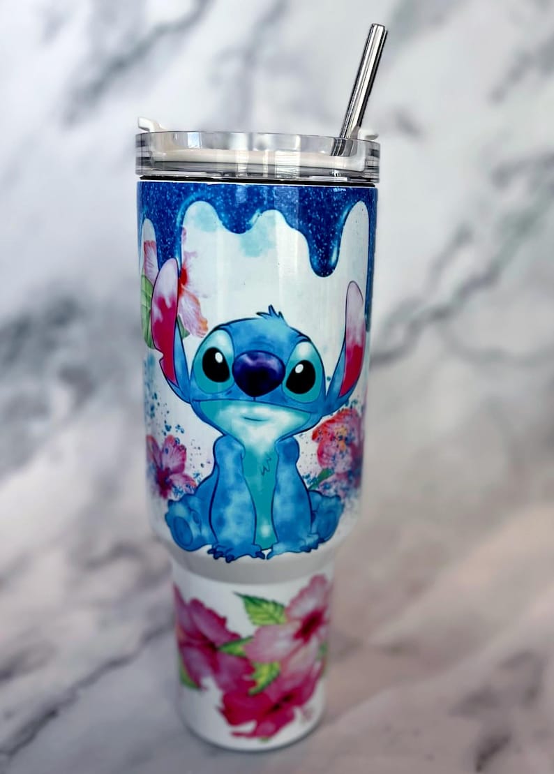 May include: A white stainless steel tumbler with a blue and white watercolor design of Stitch from Lilo & Stitch. The tumbler has a clear lid and a stainless steel straw. The design also includes pink hibiscus flowers.