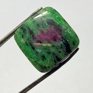 Peut inclure: Une pierre précieuse de coupe carrée, tenue par une pince, présente une base verte vibrante avec des mouchetures noires et une tache centrale rouge foncé. La surface polie reflète la lumière, révélant les motifs et les couleurs naturels de la pierre.