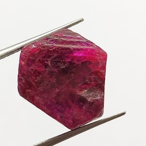 May include: A deep red ruby gemstone with a faceted, hexagonal shape. The ruby is held by tweezers against a white background.