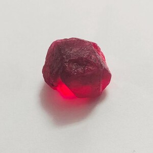 Puede incluir: Una gema de rubí rojo oscuro en bruto. El rubí tiene una superficie texturizada y una forma irregular, con una apariencia vibrante y brillante. La gema está sobre un fondo blanco liso.