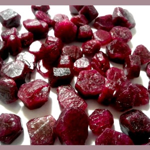 May include: A collection of raw ruby gemstones. The stones are a deep red colour, with a rough, textured surface. The gemstones are scattered across a white surface, showcasing their natural, irregular shapes.