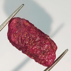Puede incluir: Una piedra preciosa de rubí rojo oscuro sin pulir. El rubí es de color rojo intenso y tiene una textura rugosa y natural.