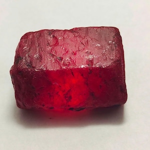 Puede incluir: Una gema de rubí en bruto, sin cortar, de color rojo intenso. La superficie de la piedra tiene un aspecto rugoso y texturizado, con imperfecciones naturales visibles. La gema es de forma rectangular y parece translúcida.