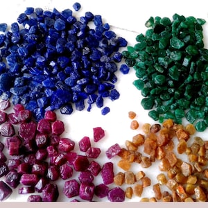 May include: A collection of loose, raw gemstones arranged in four distinct piles. The assortment includes deep blue sapphires, vibrant emerald green stones, rich magenta rubies, and warm amber-colored crystals, all featuring irregular, faceted shapes.