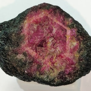 May include: A rough, dark green and black rock with a large, bright pink ruby crystal inside. The ruby crystal is partially exposed and has a rough, textured surface.