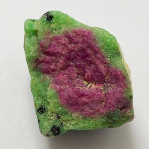 Puede incluir: Una gema en bruto con un centro magenta vibrante rodeado por un exterior verde moteado. La piedra tiene una forma natural e irregular, con pequeñas inclusiones negras dispersas por la superficie. Los colores son vivos y la textura parece áspera.