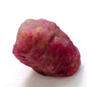 May include: A rough, pink ruby gemstone with a slightly irregular shape. The ruby has a deep, rich pink colour and is translucent.