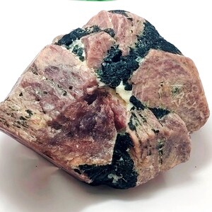 May include: A rough-cut gemstone with a mix of pink and black hues. The pink sections have a slightly translucent quality, while the black areas appear dense and opaque. The stone's surface is textured, with visible crystalline structures.