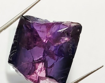 May include: A faceted purple gemstone with a rough, natural cut. The gemstone is held by tweezers against a white background.