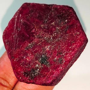 Może przedstawiać: Duży, ciemnoczerwony rubin o fasetowanym, sześciokątnym kształcie. Rubin ma szorstką, naturalną fakturę i wykazuje pewne inkluzje.