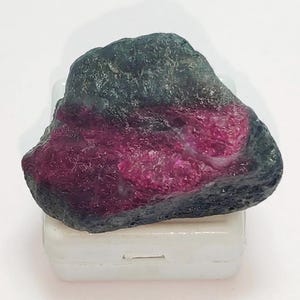 Puede incluir: Una gema en bruto, sin cortar, con un exterior gris oscuro y un interior magenta vibrante. La piedra tiene una forma irregular y descansa sobre una base rectangular blanca. El contraste entre el exterior oscuro y el interior brillante crea un efecto visual llamativo.