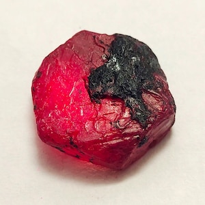 Peut inclure: Une pierre précieuse de rubis brute et non taillée. Le rubis est d'un rouge profond et vibrant, avec une surface rugueuse et texturée. Une zone noire, possiblement de la matrice, est visible sur un côté. La pierre est de forme irrégulière.