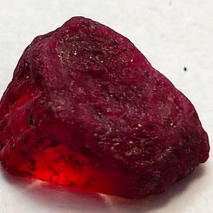 Peut inclure: Une pierre précieuse brute de rubis non taillé, d'un rouge profond. La surface est texturée avec des variations de tons, et une zone translucide brillante est visible. Ce spécimen minéral brut est une pierre précieuse naturelle.