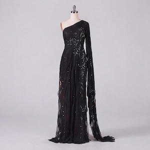 May include: A floor-length black one-shoulder gown featuring sheer fabric with intricate black sequin embellishments. The dress includes a single long, flowing sleeve and a draped bodice that extends into a full-length skirt with a side slit.