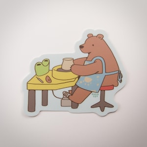 May include: A die-cut sticker featuring a brown bear wearing a light blue apron with paw prints, sitting on a red stool while working at a yellow pottery wheel table. The bear is shaping a clay pot. The text DUCK LEY is printed near the bear's leg.