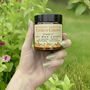 May include: A small glass jar with a black lid, labeled "earthglow emporium CITRUS GROVE Grapefruit SOY WAX CANDLE." The label includes text about the candle being all-natural, organic, and hand-poured. The jar contains four ounces of soy wax.