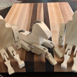 May include: Three wooden articulated figures, two human-shaped and one horse-shaped, are sitting on a wooden cutting board. The figures are made of light-colored wood and have movable joints. The horse has a brown mane.