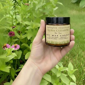 May include: A hand holds a small glass jar with a black lid. The jar contains a soy wax candle with a label that reads "Aura Cleanse Tea Tree" and "Four Ounces". The background features green foliage and pink flowers.