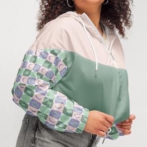 May include: A cropped windbreaker jacket with a color-block design. The top section is light pink, the middle is sage green, and the sleeves feature a repeating pattern of moons and stars in green, purple, and white. The jacket has a hood and a zipper.