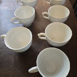 May include: A set of seven white ceramic tea cups arranged in a group. Each cup features a smooth, glossy finish and a subtle, spiraling swirl pattern embossed into the exterior surface, complete with a small, curved handle on the side.