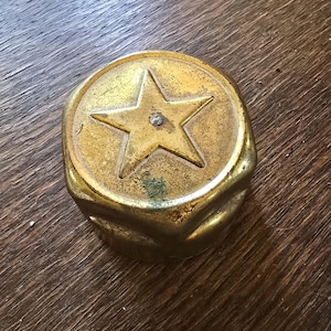 May include: A circular, metallic gold-toned cap featuring a raised five-pointed star design centered on the top surface. The object has a weathered, slightly tarnished finish with visible signs of oxidation and a small, dark spot near the star.
