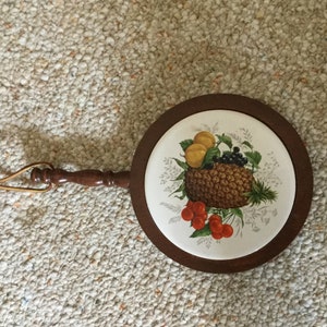 Könnte beinhalten: Ein runder Holz-Topfuntersetzer mit einem Keramik-Einsatz, der ein gemaltes Design aus Ananas, Kirschen, Zitronen und Brombeeren zeigt.