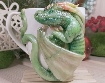 May include: A white ceramic mug with a detailed, green dragon sculpture wrapped around it. The dragon has iridescent wings and a textured body. The mug is set on a small wooden platform, with festive decorations in the background.