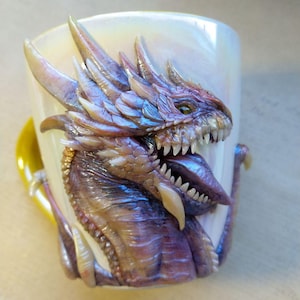 Könnte beinhalten: Eine Keramiktasse mit einem detailliert modellierten Drachenkopf. Der Drache ist lila, braun und weiß mit einem strukturierten, schuppigen Design. Die Tasse ist weiß mit einem Perlmutt-Finish.