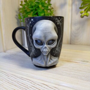 May include: A black ceramic mug with a 3D sculpted gray alien face. The alien has large eyes and a serious expression. The mug has a white background with a black galaxy design.