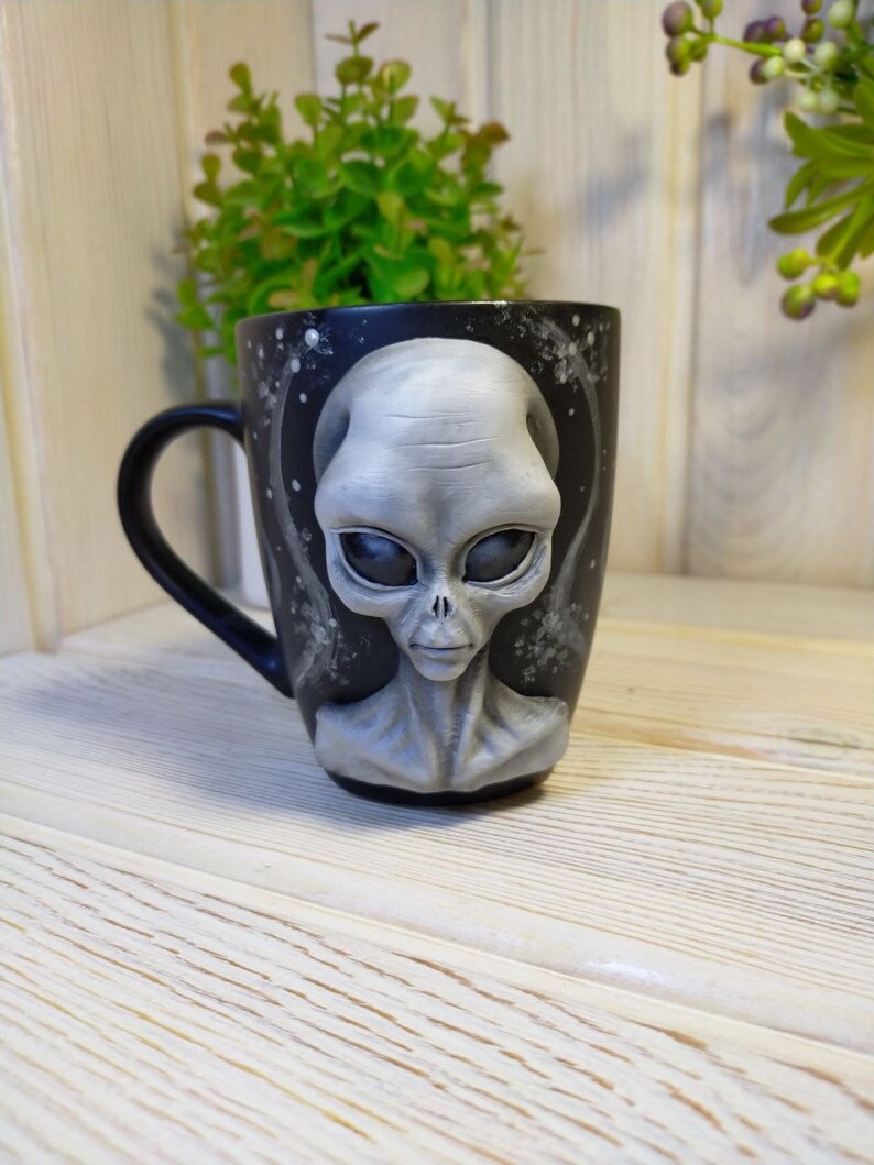 May include: A black ceramic mug with a 3D sculpted gray alien face. The alien has large eyes and a serious expression. The mug has a white background with a black galaxy design.