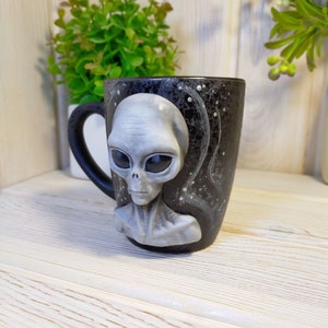 May include: A black ceramic mug with a gray alien face sculpted on the front. The mug has a white speckled design and a black handle.