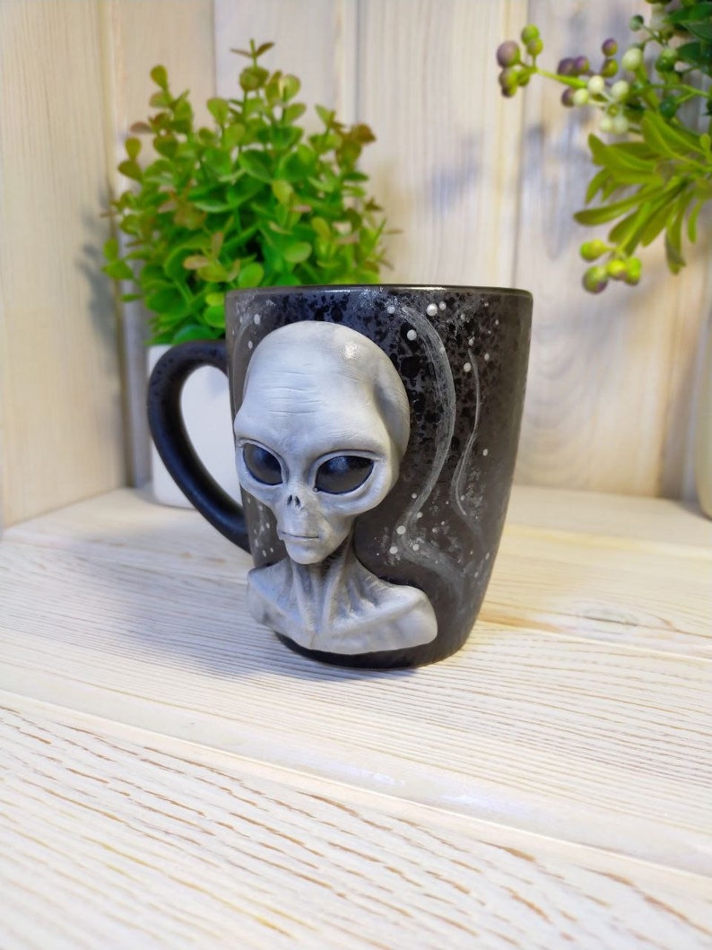 May include: A black ceramic mug with a gray alien face sculpted on the front. The mug has a white speckled design and a black handle.