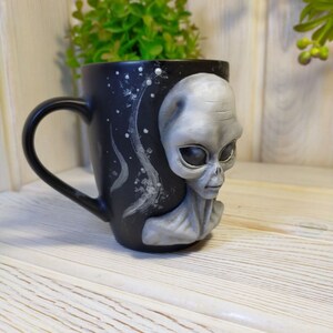 May include: Black ceramic mug with a 3D alien face sculpted on the side. The alien has gray skin, large eyes, and a small mouth. The mug has a white starry sky design on the back.