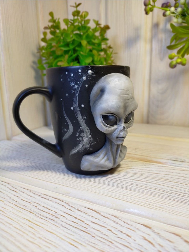 May include: Black ceramic mug with a 3D alien face sculpted on the side. The alien has gray skin, large eyes, and a small mouth. The mug has a white starry sky design on the back.