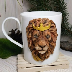 May include: White ceramic mug with a 3D sculpted lion's head wearing a gold crown. The lion's mane is brown and the eyes are brown.