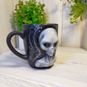 May include: A black ceramic mug with a gray alien face on the front. The mug has a white and black starry sky design.