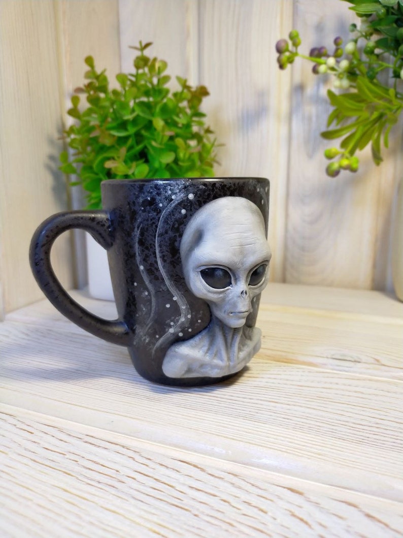 May include: A black ceramic mug with a gray alien face on the front. The mug has a white and black starry sky design.