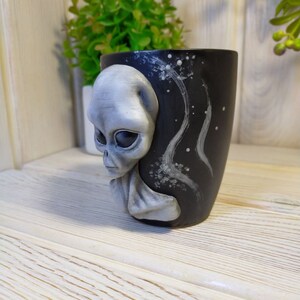 May include: A black ceramic mug with a grey alien face sculpted on the side. The mug has a white and grey galaxy design on the back.