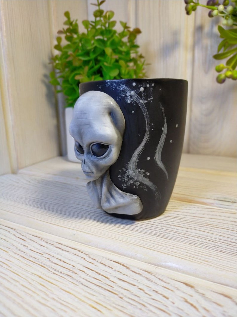 May include: A black ceramic mug with a grey alien face sculpted on the side. The mug has a white and grey galaxy design on the back.