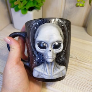 May include: A black ceramic mug with a gray alien face sculpted on the front. The alien has large eyes and a serious expression. The mug is decorated with white dots on a black background, resembling stars.
