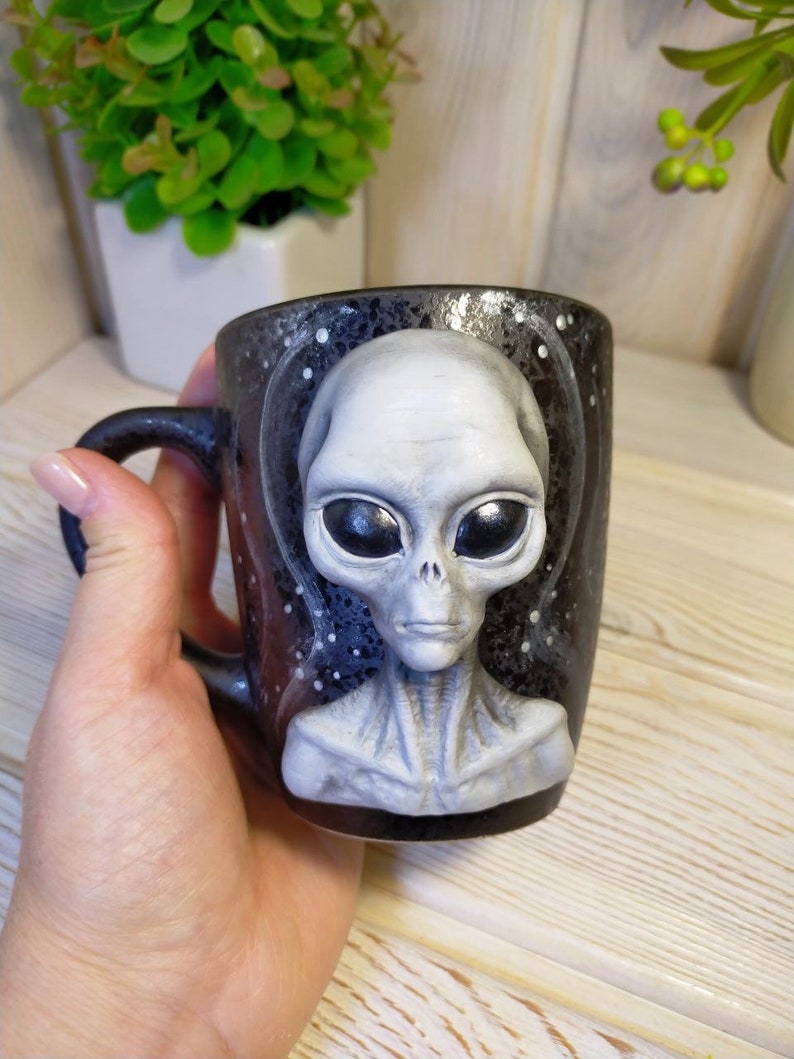 May include: A black ceramic mug with a gray alien face sculpted on the front. The alien has large eyes and a serious expression. The mug is decorated with white dots on a black background, resembling stars.