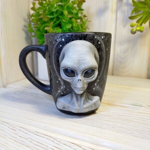 May include: Black ceramic mug with a 3D sculpted gray alien face. The mug has a black speckled glaze and a white background.